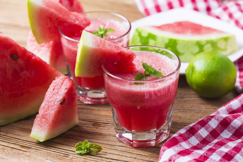 Suco de melancia com hortelã servido em copo de vidro em cima de mesa de madeira ao redor está pedaços de melancia e folhas de hortelã e um limão
