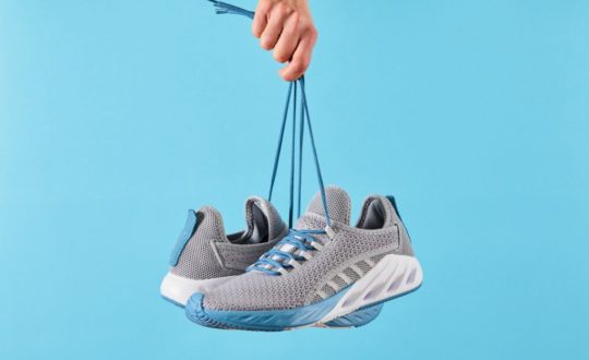 Imagem de uma mão segurando um tenis cinza com solado branco e detalhes em azul, em fundo azul-claro