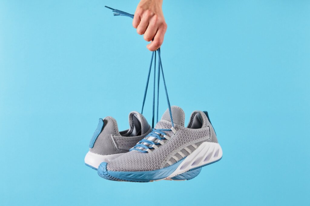 Imagem de uma mão segurando um tenis cinza com solado branco e detalhes em azul, em fundo azul-claro