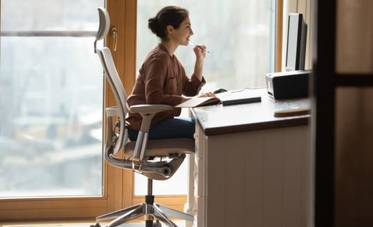 Mulher de cabelo preso sentada em cadeira ergonômica e de frente para monitor de computador