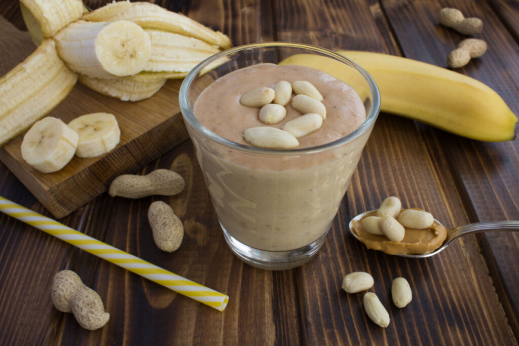 Copo de vitamina de banana com pasta de amendoim ao lado de amendoins, bananas cortadas e uma inteira 