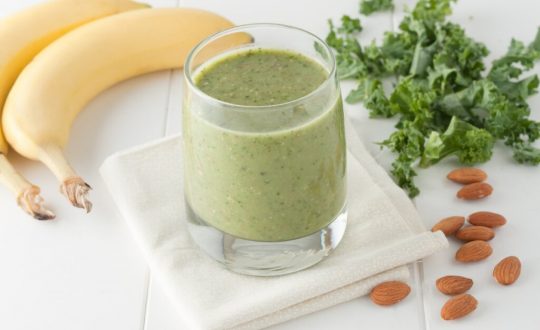 Copo de vidro com vitamina de couve com banana e amêndoas em cima de um pano branco em uma mesa de madeira branca