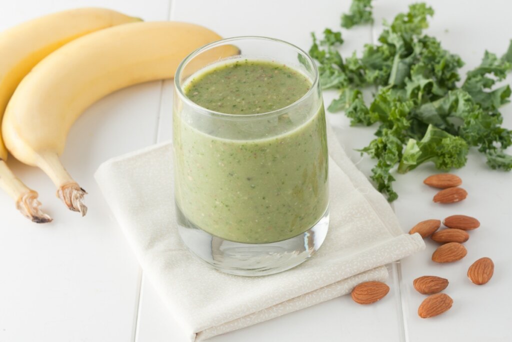 Copo de vidro com vitamina de couve com banana e amêndoas em cima de um pano branco em uma mesa de madeira branca