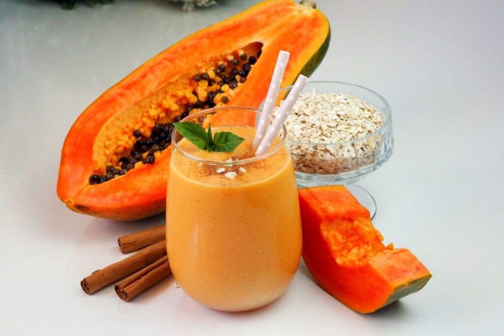 Vitamina de mamão com aveia e canela servido em copo de vidro em cima de mesa de mármore branco, ao lado está metade de um mamão, uma tigela com aveia e canela em pau 